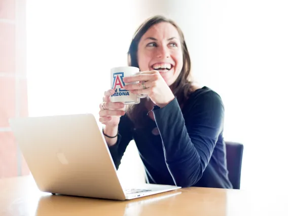 Distance learner smiling while holding their coffee mug and working at a laptop.